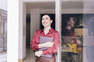 Smiling female business professional looking away while holding laptop at glass door in home - KMKF01577