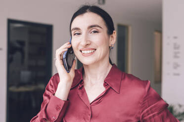 Lächelnde Geschäftsfrau, die zu Hause mit ihrem Smartphone telefoniert - KMKF01578