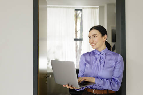 Lächelnde Geschäftsfrau, die ihren Blick abwendet, während sie einen Laptop im Heimbüro hält - KMKF01588