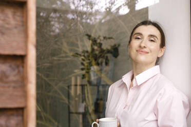 Smiling female business professional with coffee mug standing against wall at home - KMKF01599