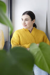 Smiling businesswoman in warm clothing looking away while sitting against wall at home - KMKF01602