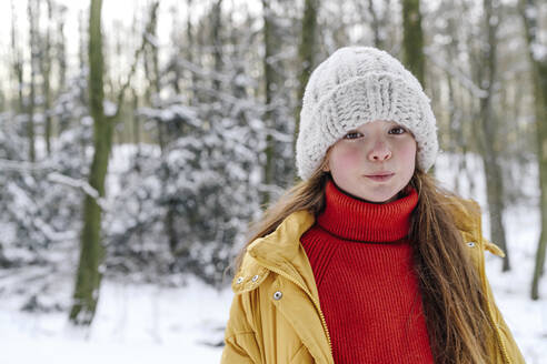 Girl wearing knit hat standing against trees during snow - OGF00931