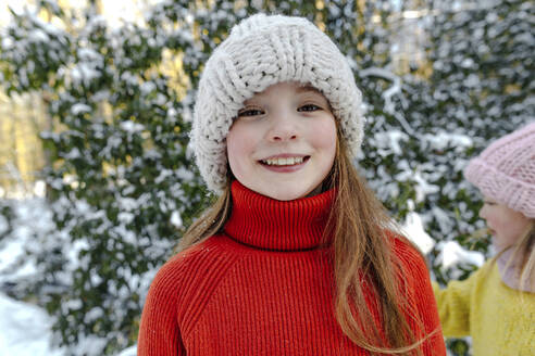 Smiling girl in knit hat standing against trees during snow - OGF00932