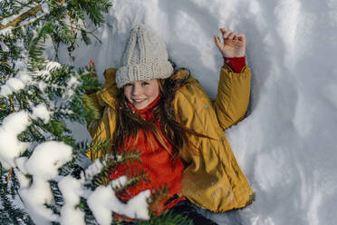 Lächelndes Mädchen in gelber Jacke liegt im Winter unter einem Baum im Schnee - OGF00935