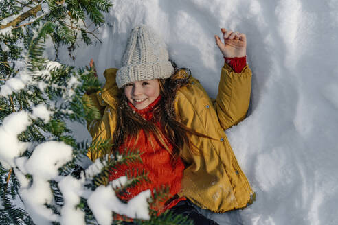 Smiling girl in yellow jacket lying on snow under tree during winter - OGF00935