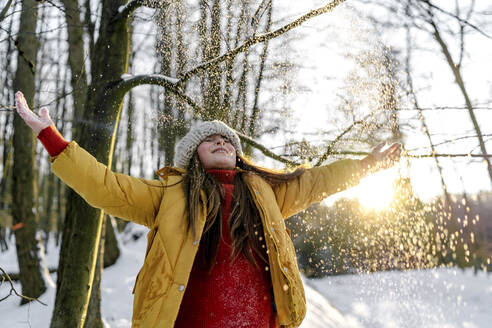 Girl in warm clothing standing with eyes closed under tree during snowfall - OGF00936