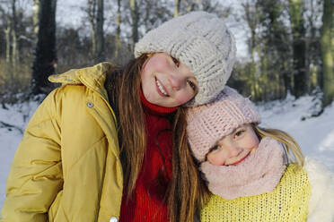 Smiling sisters in warm clothing standing together against trees during snow - OGF00937