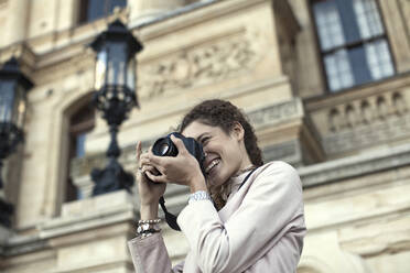 Junge Frau, die mit einer Digitalkamera gegen ein Gebäude fotografiert - AJOF01232