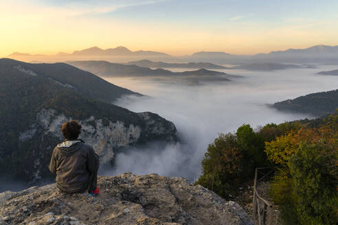 Male tourist sitting at cliff watching sunrise at Furlo Gorge, Marche, Italy - LOMF01237