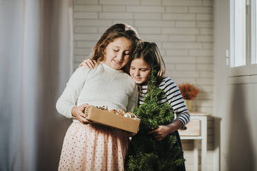 Mädchen mit Kiefer Weihnachten umarmt Schwester hält Weihnachtsschmuck Box zu Hause - MIMFF00578