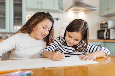 Mädchen zeichnet auf Papier, während sie mit ihrer Schwester am Esstisch in der Küche sitzt - MIMFF00582