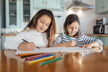 Sisters coloring with colored pencil on paper while sitting at dining table in kitchen - MIMFF00583