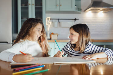 Lächelnde Mädchen schauen sich beim Malen mit Buntstiften auf Papier an, während sie am Esstisch in der Küche sitzen - MIMFF00584