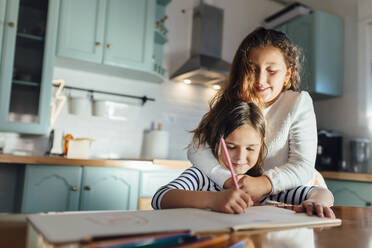 Girl coloring on paper while sister hugging from behind in kitchen - MIMFF00585