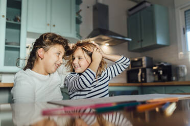 Cheerful girl playing with each other hair while sitting at dining table in kitchen - MIMFF00586