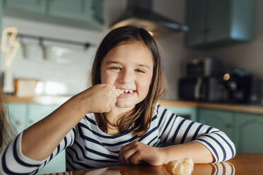 Smiling girl eating tangerine while sitting at dining table in kitchen - MIMFF00588