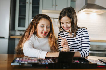 Fröhliche Schwestern spielen mit Make-up-Palette, während sie am Esstisch in der Küche sitzen - MIMFF00597