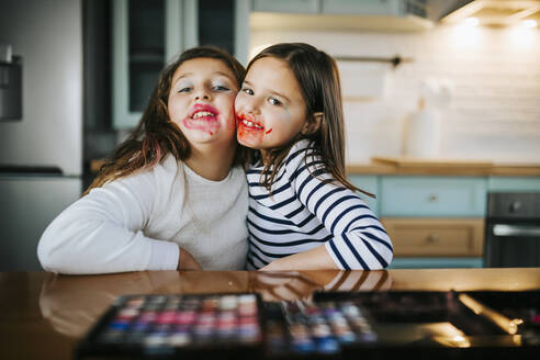 Girls with makeup on face embracing each other while sitting at dining table - MIMFF00598