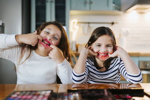 Playful girls with makeup on face teasing while sitting at dining table in domestic kitchen - MIMFF00599