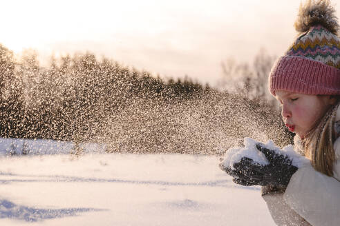 Playful girl in warm clothing blowing snow during winter - EYAF01524