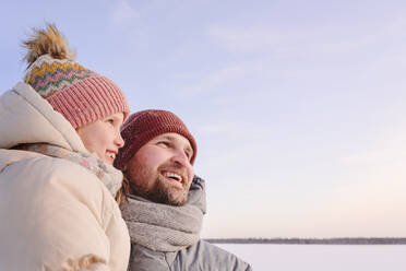 Vater und Tochter in warmer Kleidung schauen bei Sonnenuntergang weg - EYAF01532