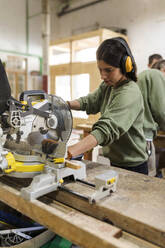 Schreinerin mit Ohrenschützer bei der Holzbearbeitung an einer Werkbank in der Industrie - VABF04248