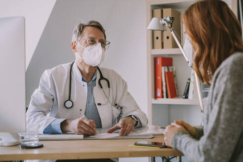 Male general practitioner with face mask looking at patient while sitting at doctor's office - MFF07492