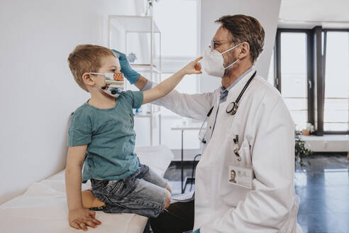 Männlicher Kinderarzt mit Gesichtsschutzmaske, der einen Jungen im medizinischen Untersuchungsraum untersucht - MFF07496