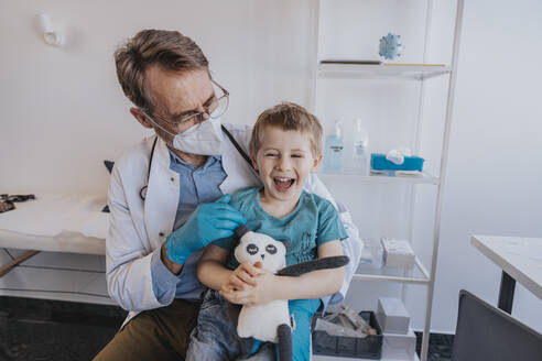 Male pediatrician looking at cheerful boy with toy while sitting at medical examination room - MFF07502