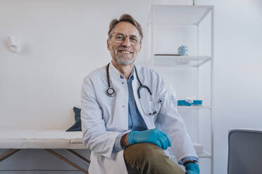 General practitioner with protective glove smiling while sitting in clinic - MFF07504