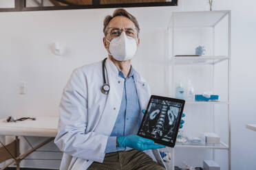 Arzt mit Gesichtsmaske zeigt Röntgenbild auf digitalem Tablet, während er in der Klinik sitzt - MFF07506