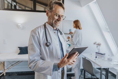 Allgemeinmediziner, der ein digitales Tablet benutzt, während er mit einem Mitarbeiter im Hintergrund in einer Klinik steht - MFF07521