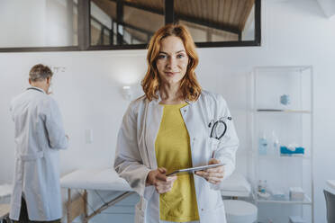 Lächelnde Ärztin, die ein digitales Tablet hält, während sie mit einem Mitarbeiter im Hintergrund in einer Klinik steht - MFF07522