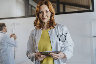 Eine Ärztin hält ein digitales Tablet, während sie mit einem Mitarbeiter im Hintergrund in einer Klinik steht - MFF07524