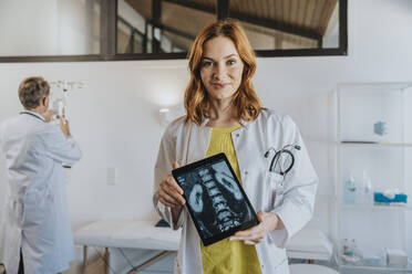 Arzt zeigt Röntgenbild auf digitalem Tablet, während er mit einem Mitarbeiter im Hintergrund in einer Klinik steht - MFF07529