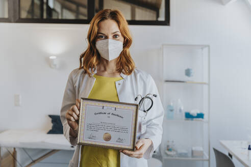 Doctor with protective face mask holding certificate frame while standing at clinic - MFF07530