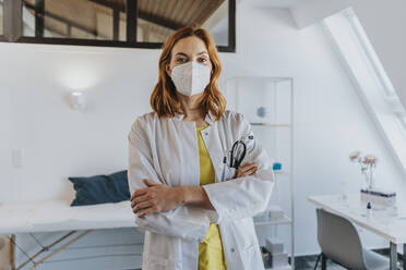 Confident healthcare worker wearing protective face mask standing with arms crossed at clinic - MFF07533