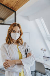 Female doctor wearing protective face mask standing with arms crossed at clinic - MFF07534