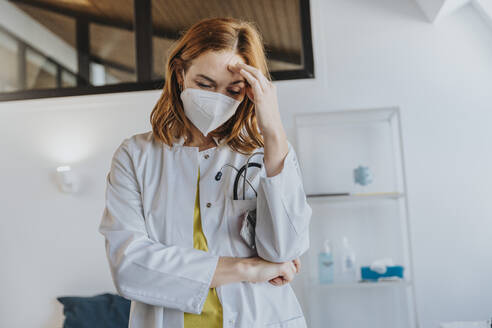 Doctor wearing protective face mask standing with head in hands at clinic - MFF07536