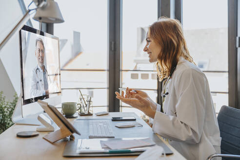 Healthcare worker talking to coworker on video call over computer while sitting at office - MFF07549