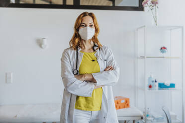 Confident female doctor wearing protective face mask standing with arms crossed at clinic - MFF07578