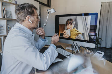 Älterer Arzt im Gespräch mit einem Patienten per Videoanruf über einen Computer, während er in der Arztpraxis sitzt - MFF07583