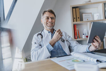 Lächelnder männlicher Arzt mit digitalem Tablet, der am Schreibtisch in einer Arztpraxis sitzt - MFF07598