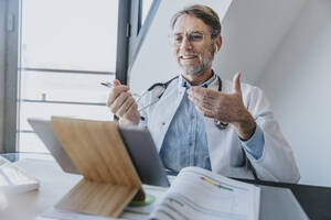 Lächelnder Arzt, der einen Videoanruf über ein digitales Tablet führt, während er in einer Klinik sitzt - MFF07599