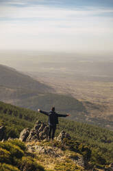 Male tourist with arms outstretched standing on rock during vacations at Somosierra, Madrid, Spain - RSGF00579