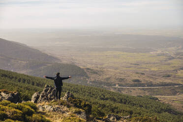 Mann mit ausgestreckten Armen, der auf einem Felsen in Somosierra, Madrid, Spanien, steht und die Aussicht betrachtet - RSGF00580