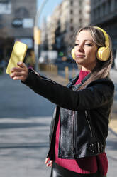 Frau in Lederjacke, die ein Selfie mit ihrem Mobiltelefon macht, während sie auf der Straße steht - AGOF00074