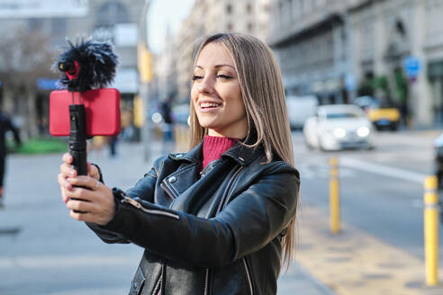 Lächelnde Frau, die auf der Straße einen Vlog über ihr Handy führt - AGOF00075