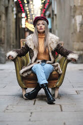 Woman with fur jacket sitting on armchair at alley - AGOF00082
