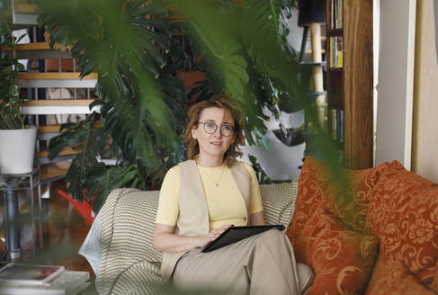 Rothaarige Geschäftsfrau mit digitalem Tablet, die zu Hause auf dem Sofa sitzt - KMKF01620